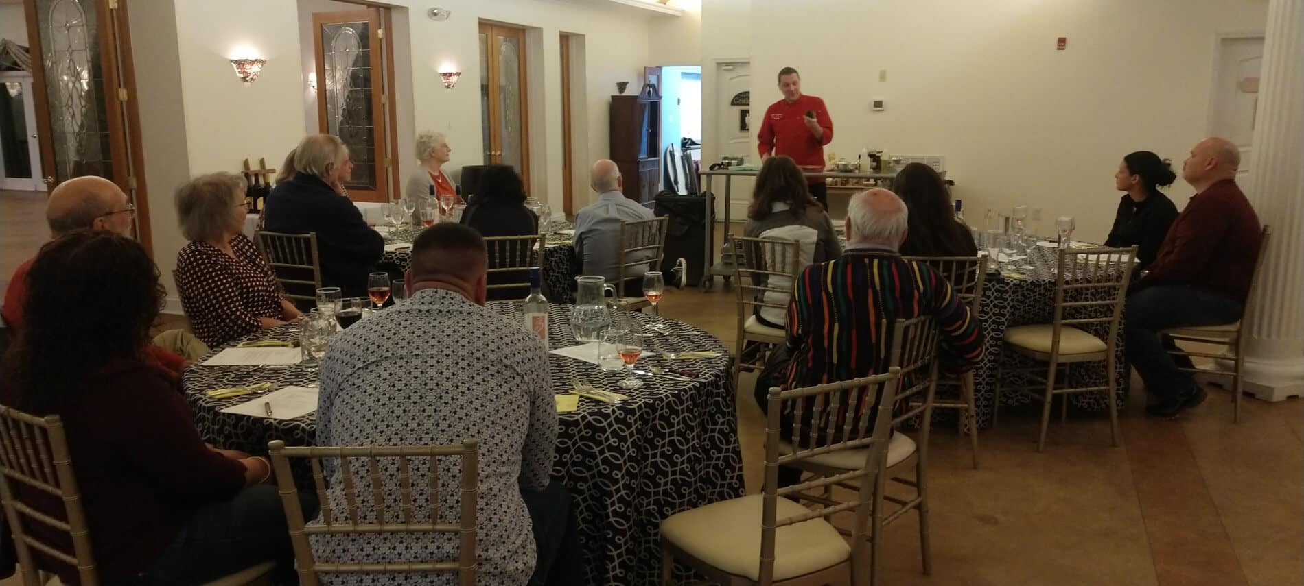 Chef Matt in red shirt teaching class of attendees sitting in cream room with round tables covered with cloths and cushioned chairs.