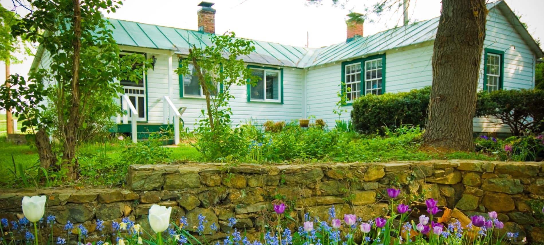 Exterior view of cottage painted white with green trim surrounded by green grass and shrubs, a stone retaining wall, and white, purple, and blue flowers