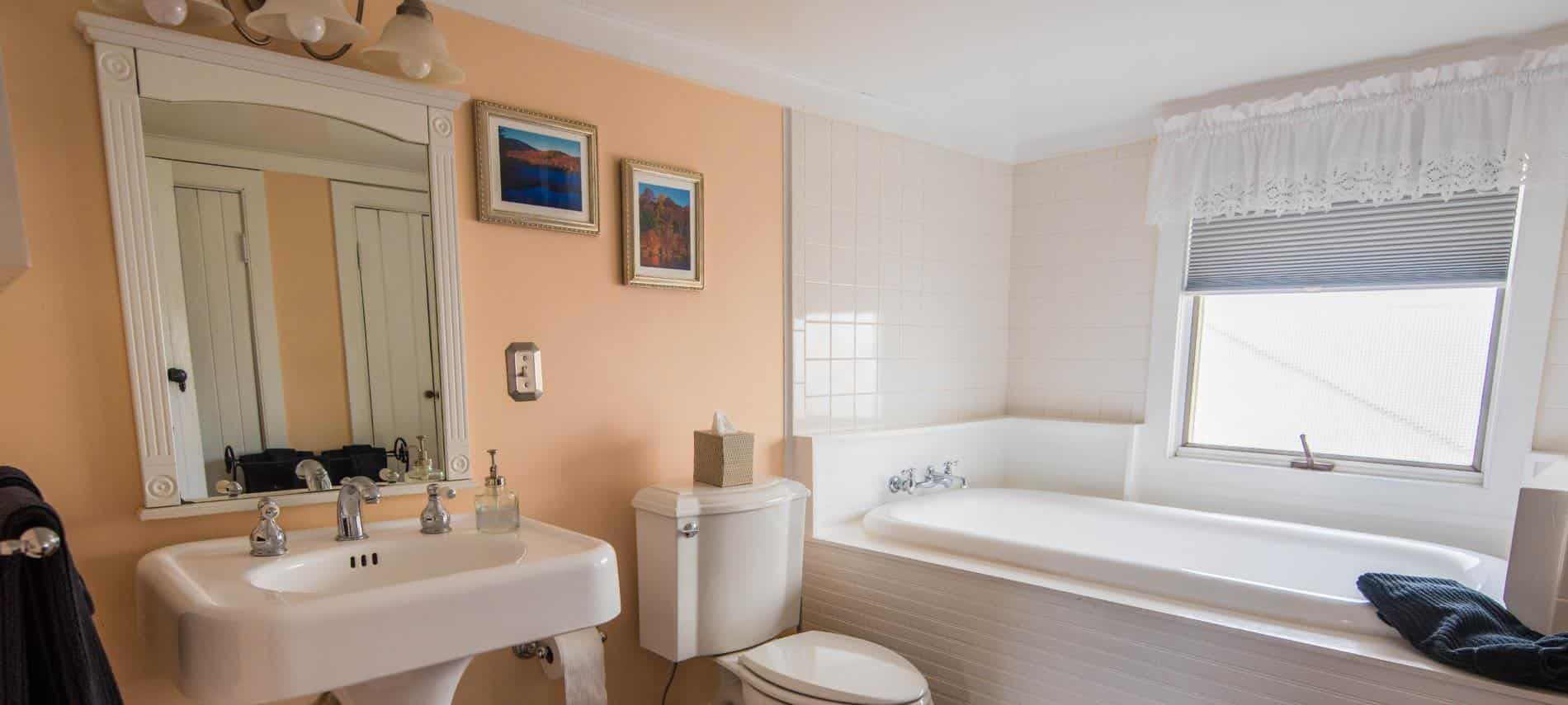 Bathroom with white tiled walls around tub, light peach colored accent wall, white pedestal sink, and white wooden framed mirror