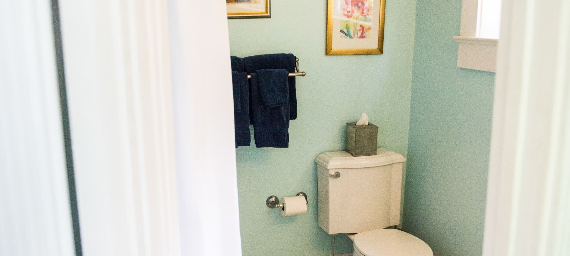 Bathroom with light mint colored walls, white toilet, white shower curtain, and navy towels