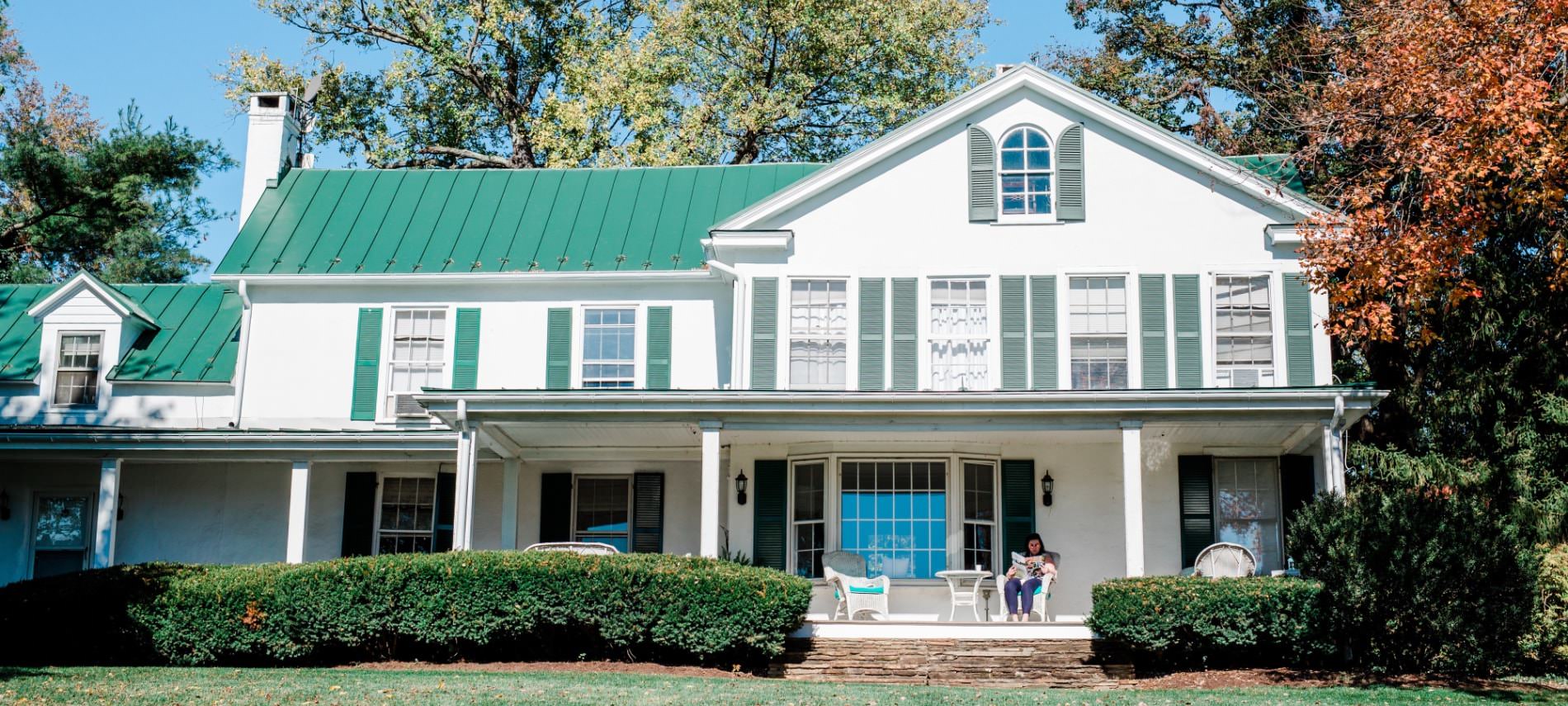 Exterior view of property painted white with green shutters and green roof surrounded by green shrubs and trees