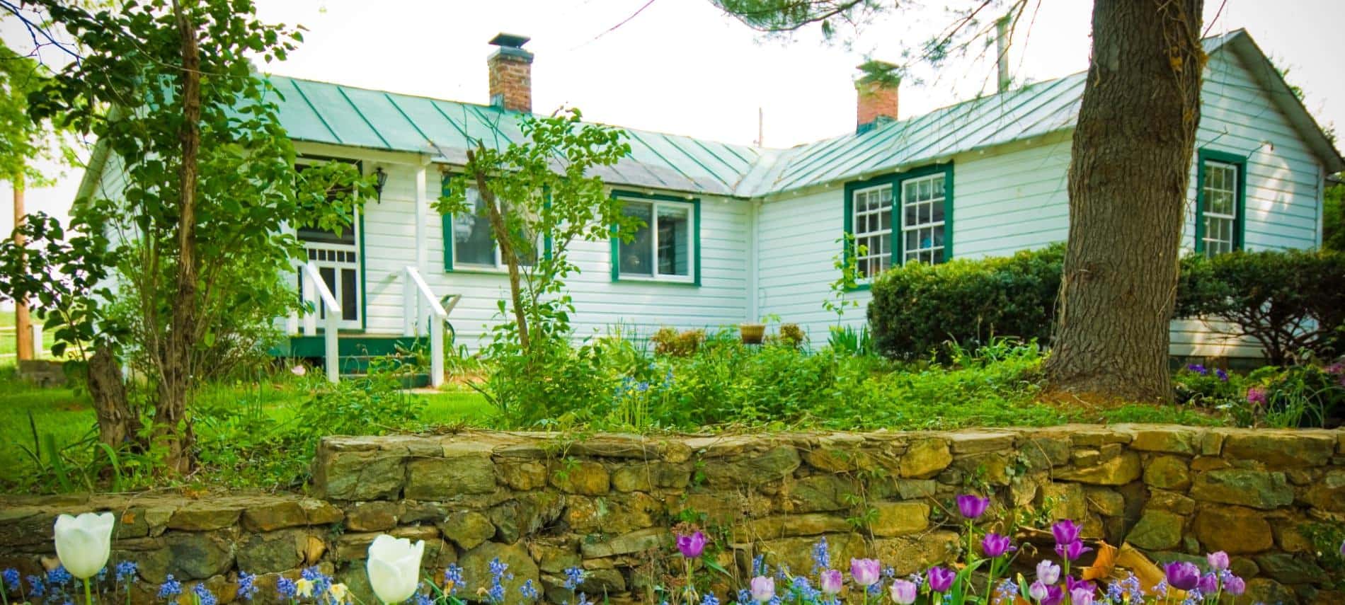Exterior view of cottage painted white with green trim surrounded by green grass and shrubs, a stone retaining wall, and white, purple, and blue flowers