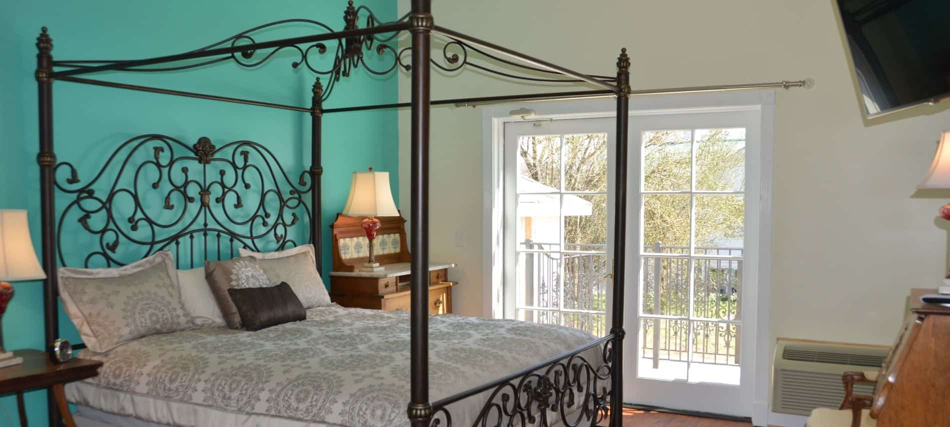 Bedroom with rod iron four-post canopy bed with light tan bedding, hardwood flooring, and a dark turquoise accent wall
