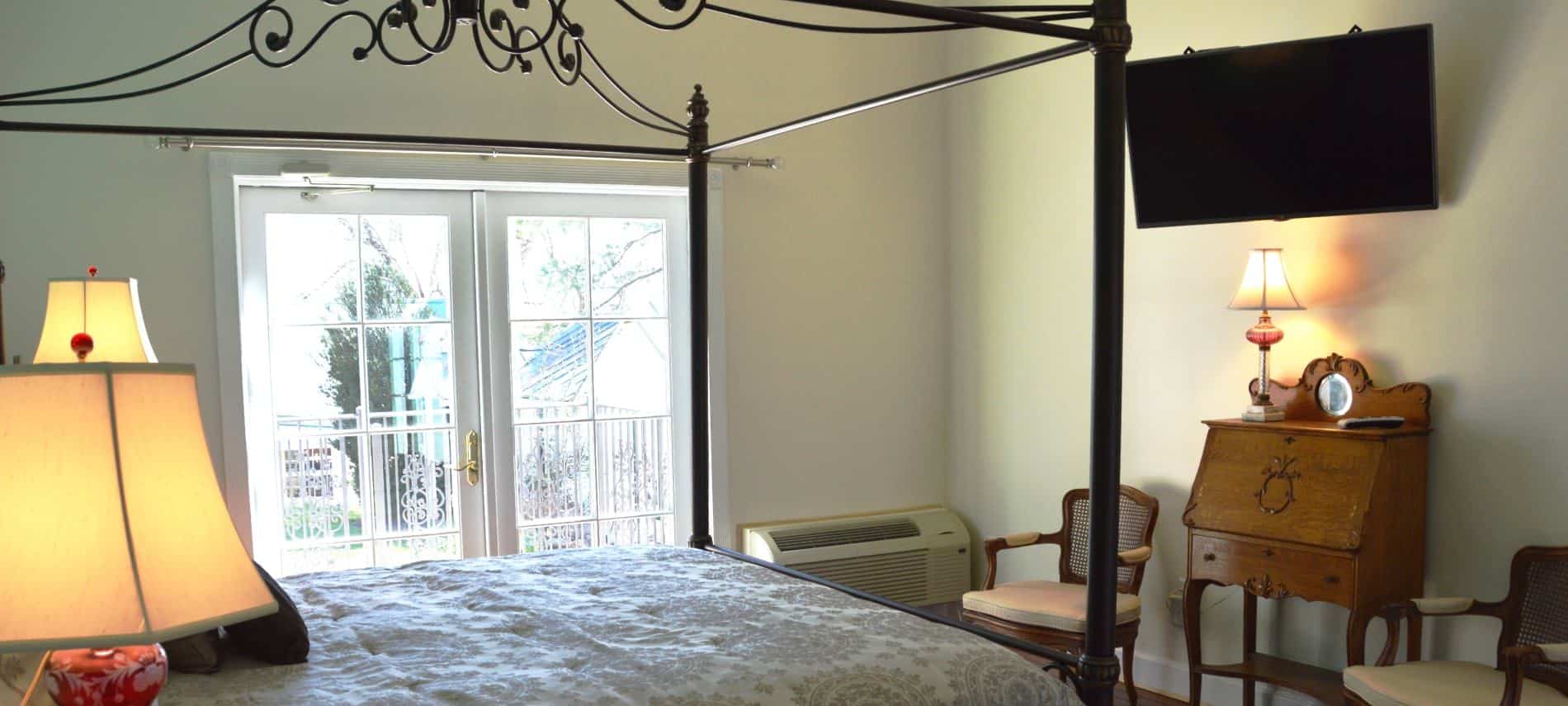 Bedroom with rod iron four-post canopy bed with light colored bedding, hardwood flooring, and flat-screen tv mounted on wall