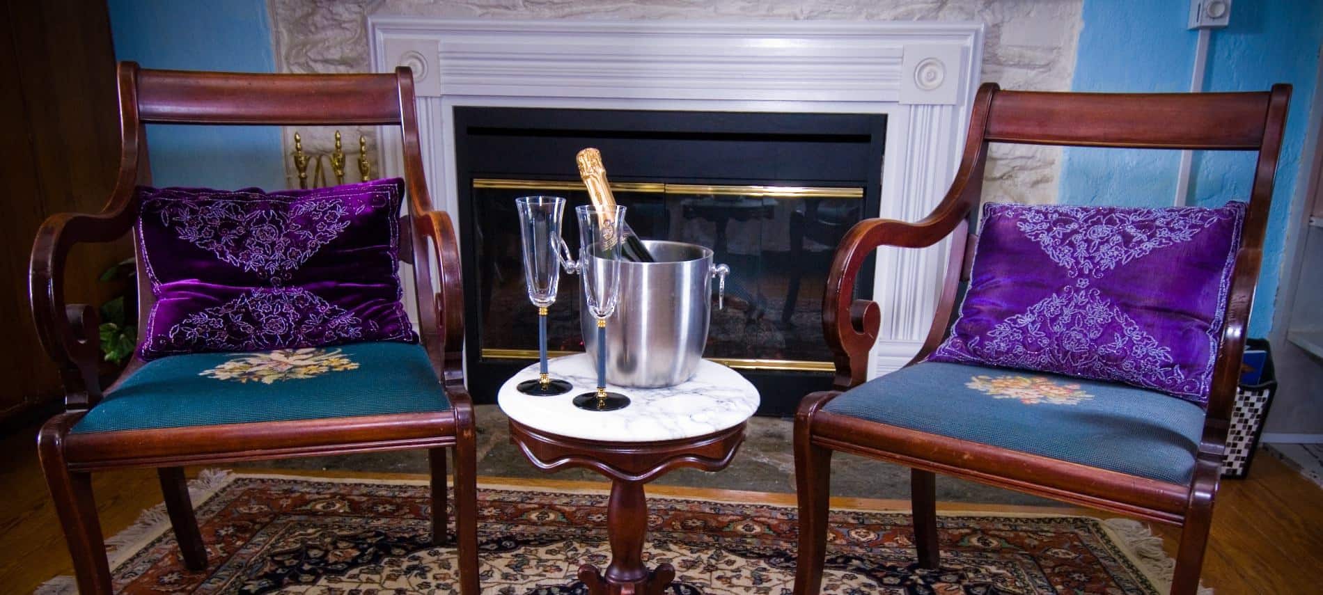 Two dark wooden chairs with dark blue upholstered seat with purple pillows next to a stone fireplace and small table with champagne bottle in ice bucket and two glasses
