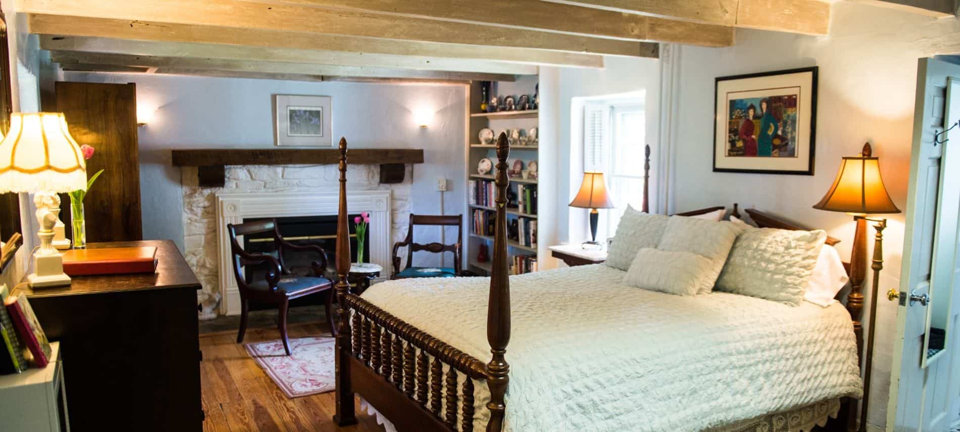 Bedroom with white bedding, light colored walls, hardwood flooring, fireplace with white stone and wooden mantel, and a sitting area
