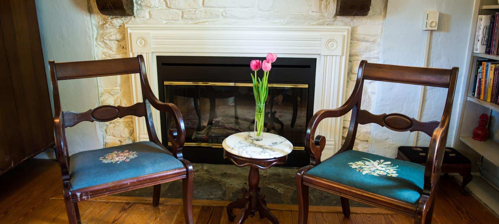 Two dark wooden chairs with dark blue upholstered seat next to a stone fireplace and small table with glass vase and pink tulips