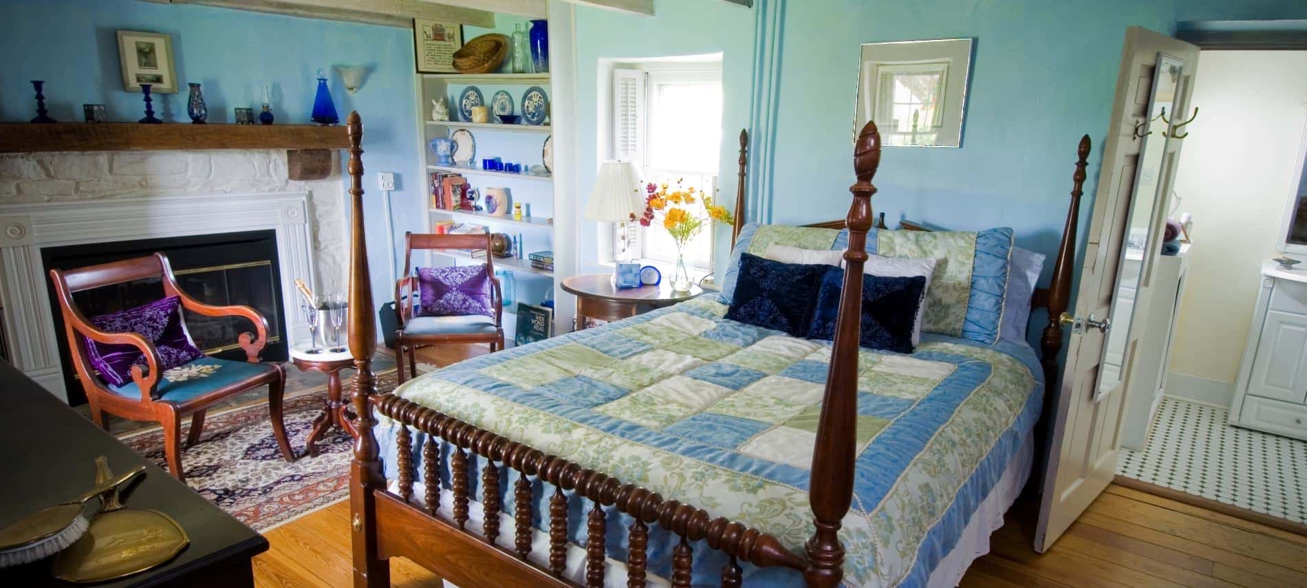 Bedroom with turquoise, sage green, and white bedding, light turquoise walls, hardwood flooring, fireplace with white stone and wooden mantel, and a sitting area
