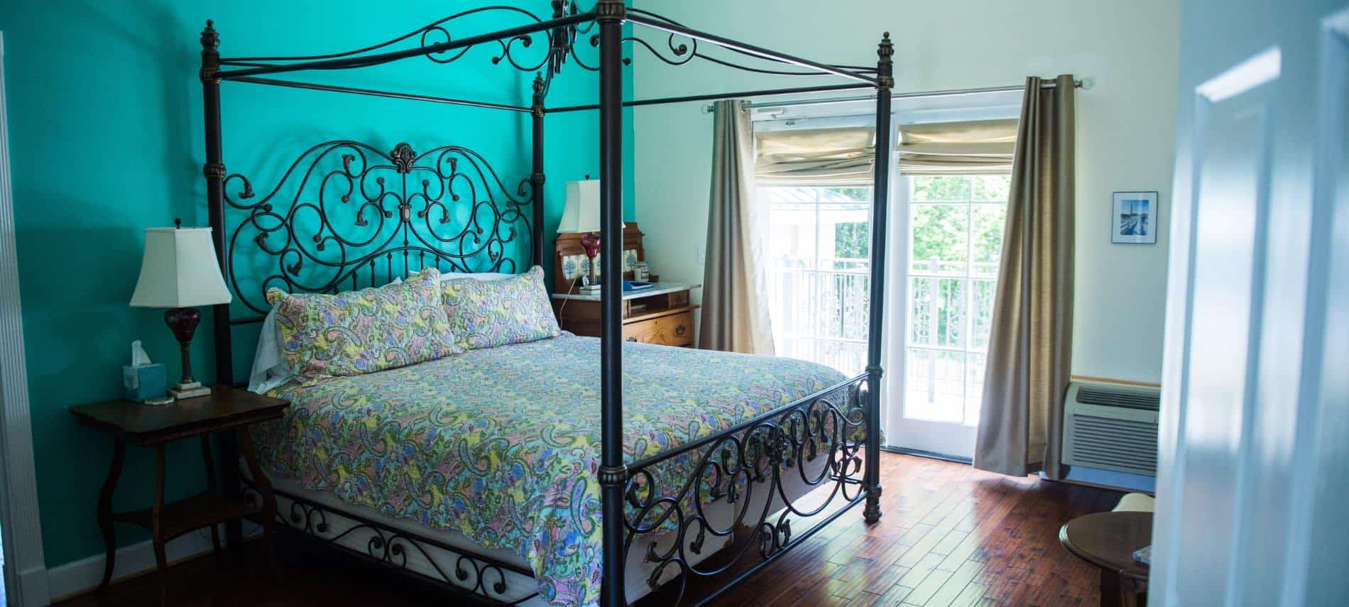 Bedroom with rod iron four-post canopy bed with multicolor paisley bedding, hardwood flooring, and a dark turquoise accent wall