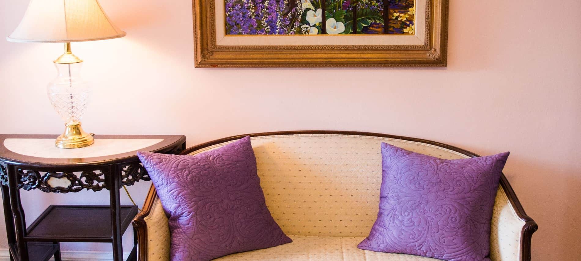 Antique wooden and cream upholstered love seat with two quilted purple pillows next to dark wooden side table with glass and brass lamp