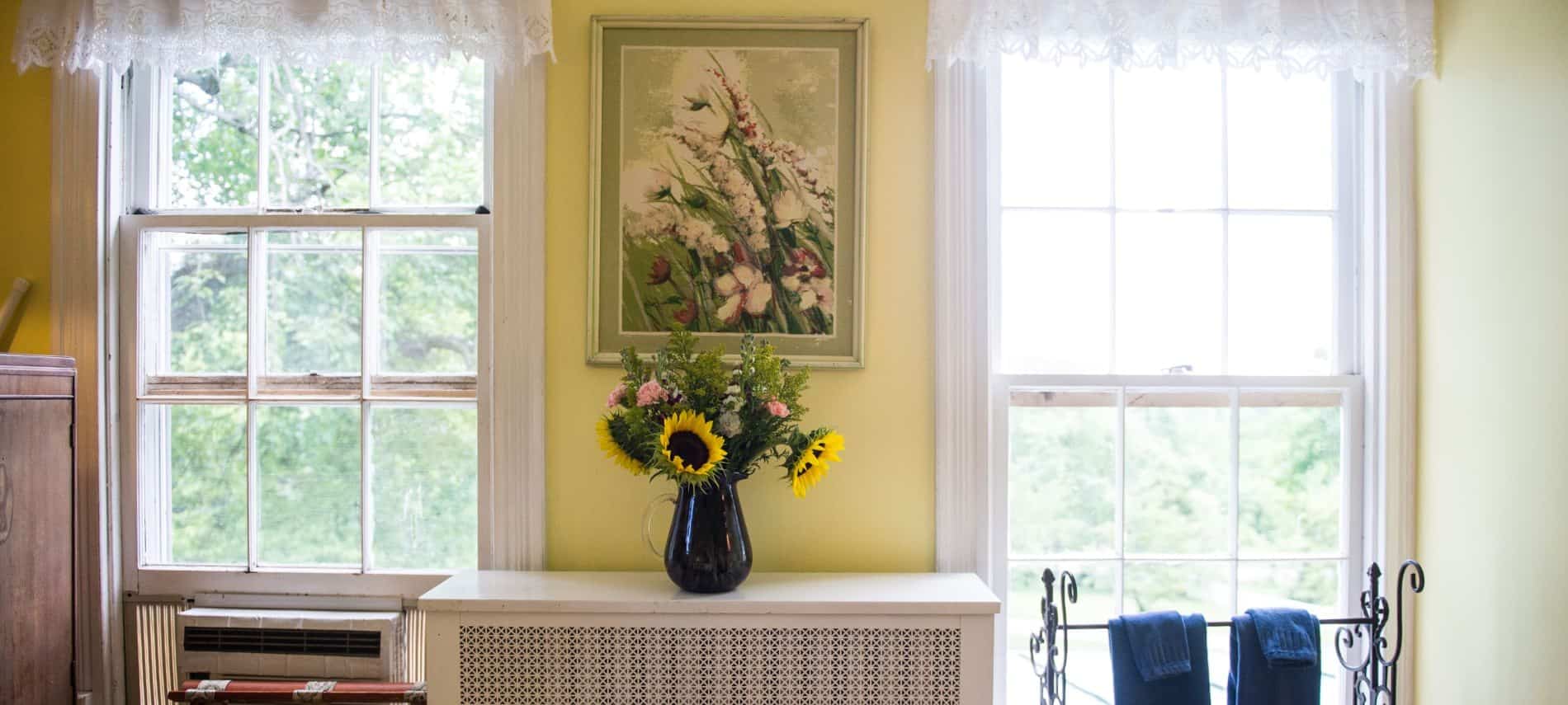 Bedroom with yellow walls, two windows with white trim and white lacey curtains, and a dark glass vase with large yellow sunflowers and small pink flowers