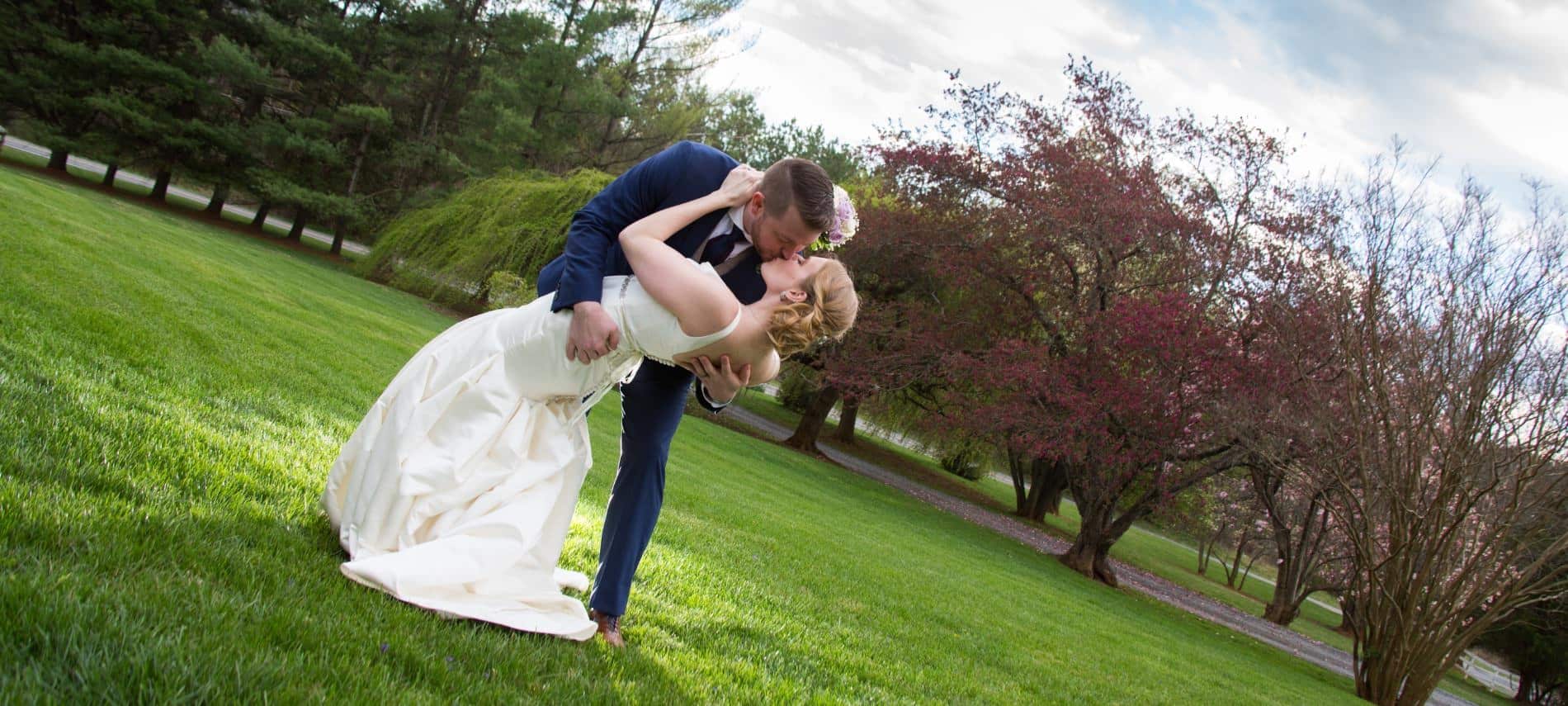Bride in white dress and groom in dark blue suit kissing in the grass with trees in the background