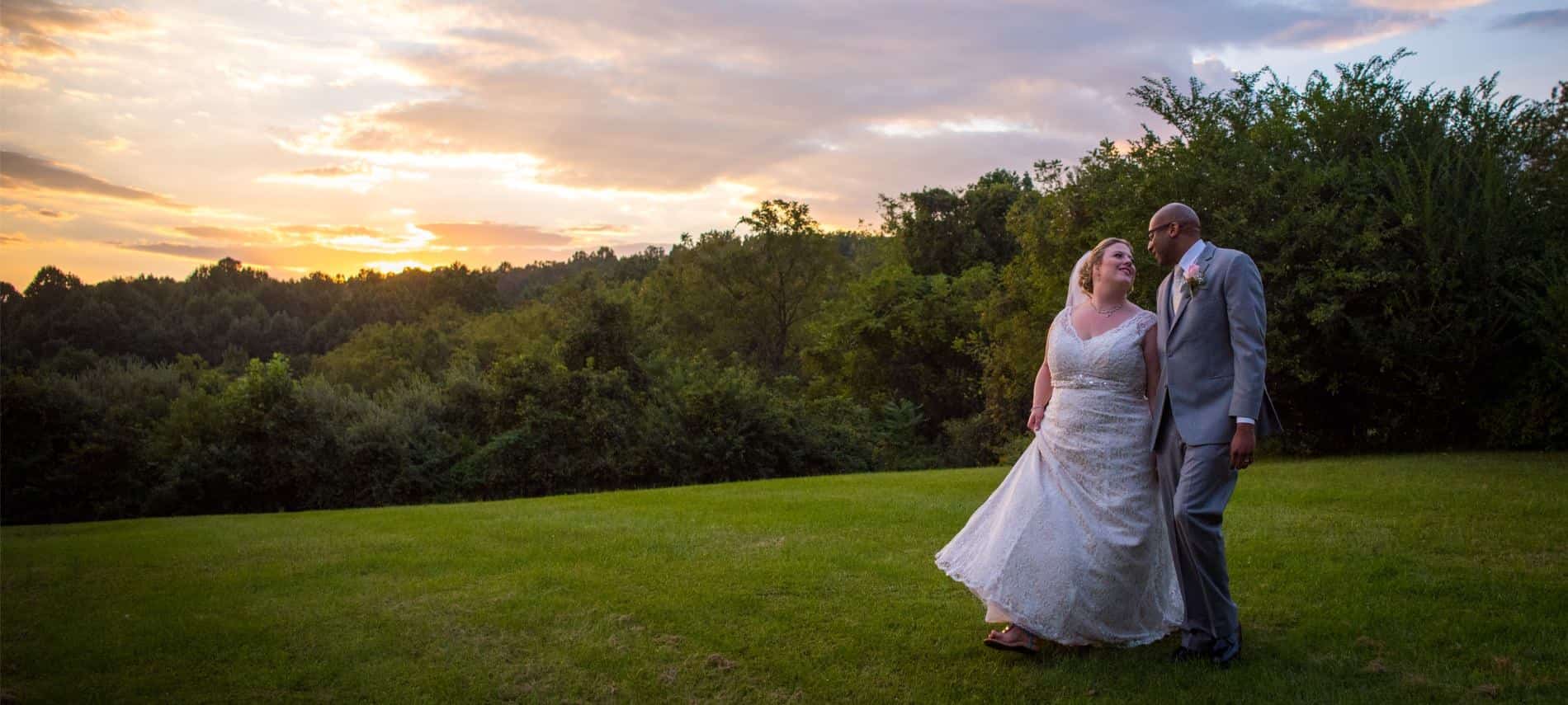 Bride in white dress and groom in gray suit standing in the grass with trees in the background at sunset