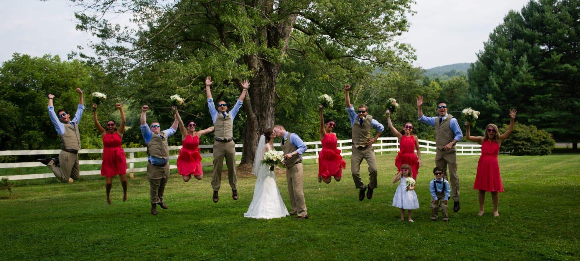 Wedding party dressed in white, tan, blue, and red with the bride and groom kissing and the rest jumping up in the air