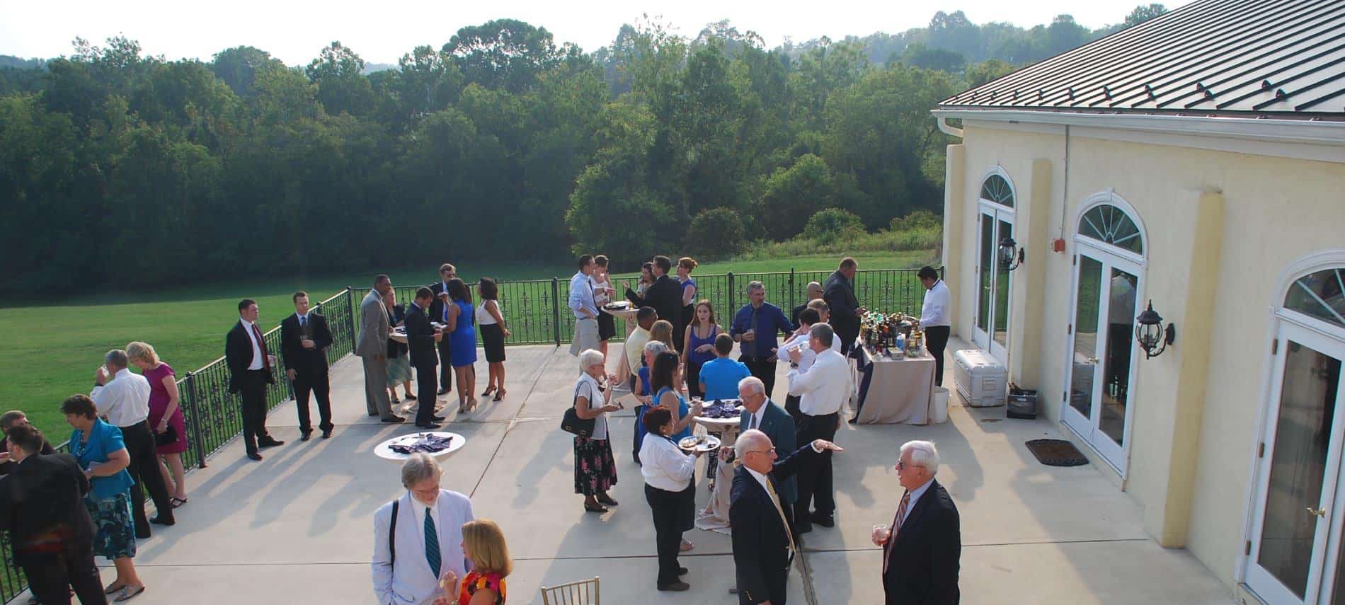 Concrete patio set up for a cocktail reception with guests eating, drinking, and talking with large green trees in the background