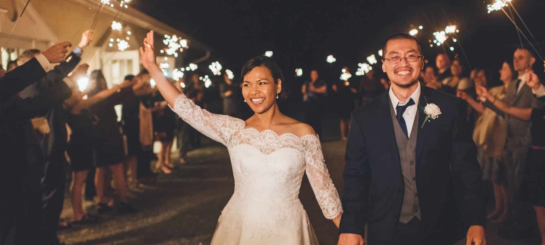 Bride in white dress and groom in black suit walking between people with sparklers