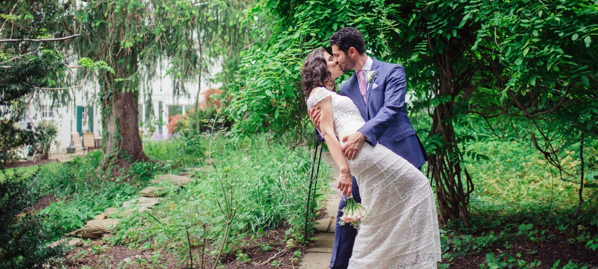 Bride with white wedding dress and groom with dark purple suit standing on stone path between large trees