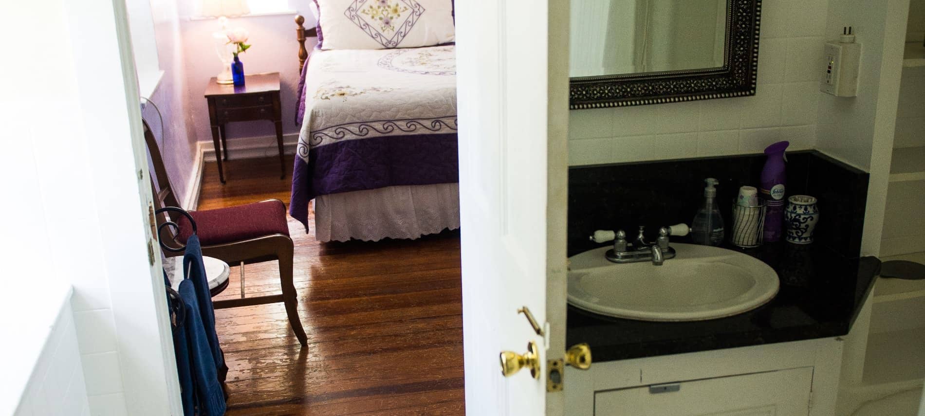 Bedroom with dark wood furniture, dark purple and white bedding, hardwood floors, and light purple walls and view of white bathroom sink and mirror