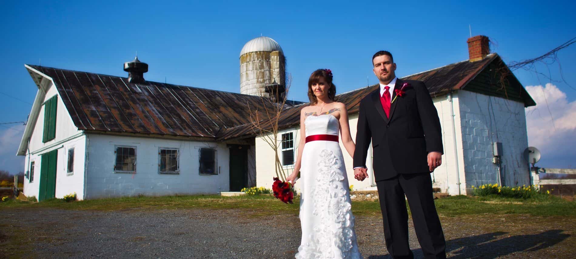 Bride in white dress and groom in black suit standing in front of old barn and silo