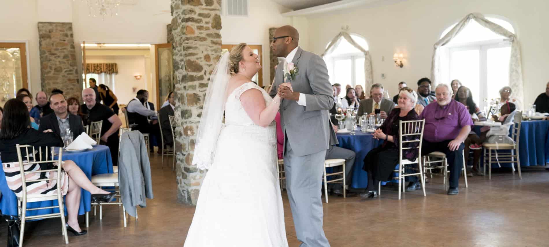 Bride in white dress and groom in gray suit dancing with people in the background watching the couple