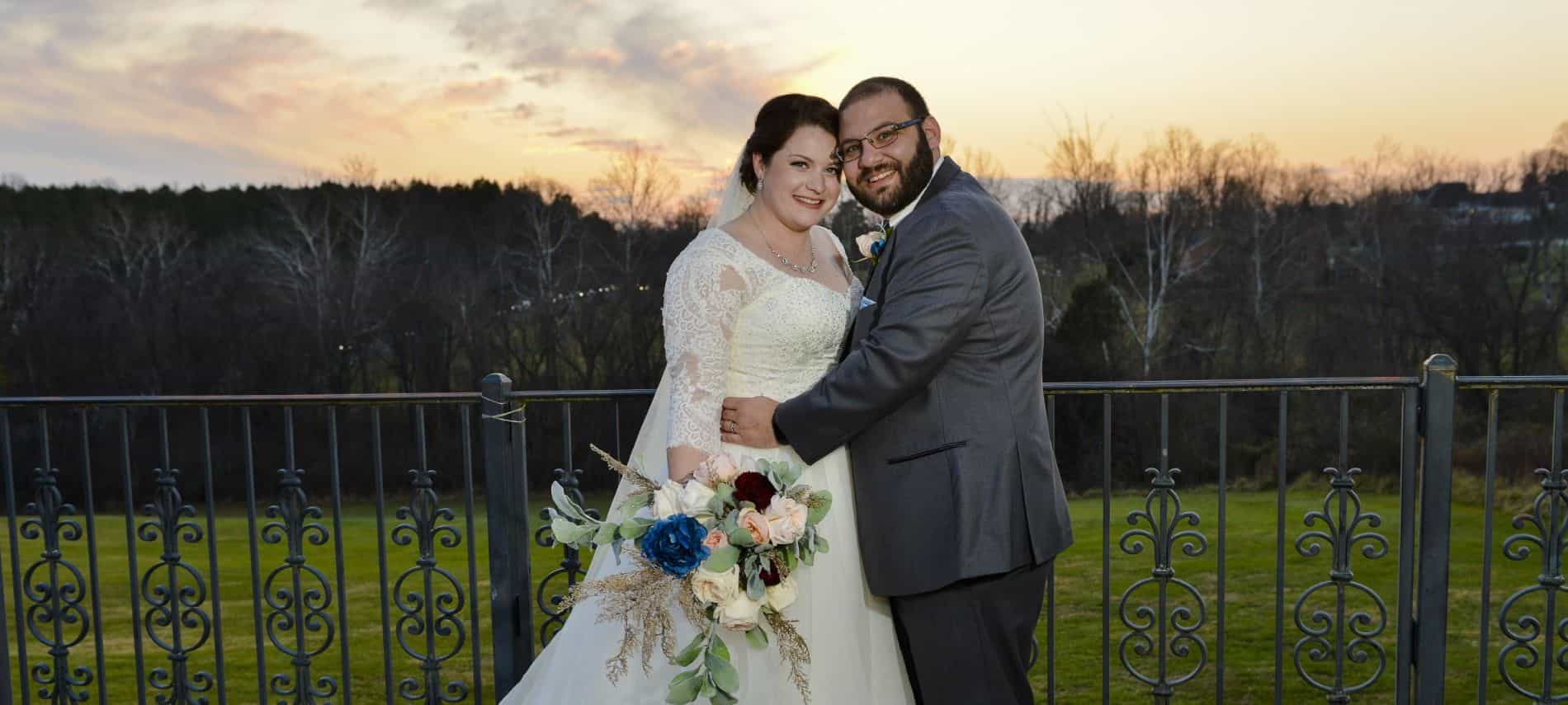 Bride in cream wedding dress and groom in dark gray suit standing near rod iron fence with sunset in background