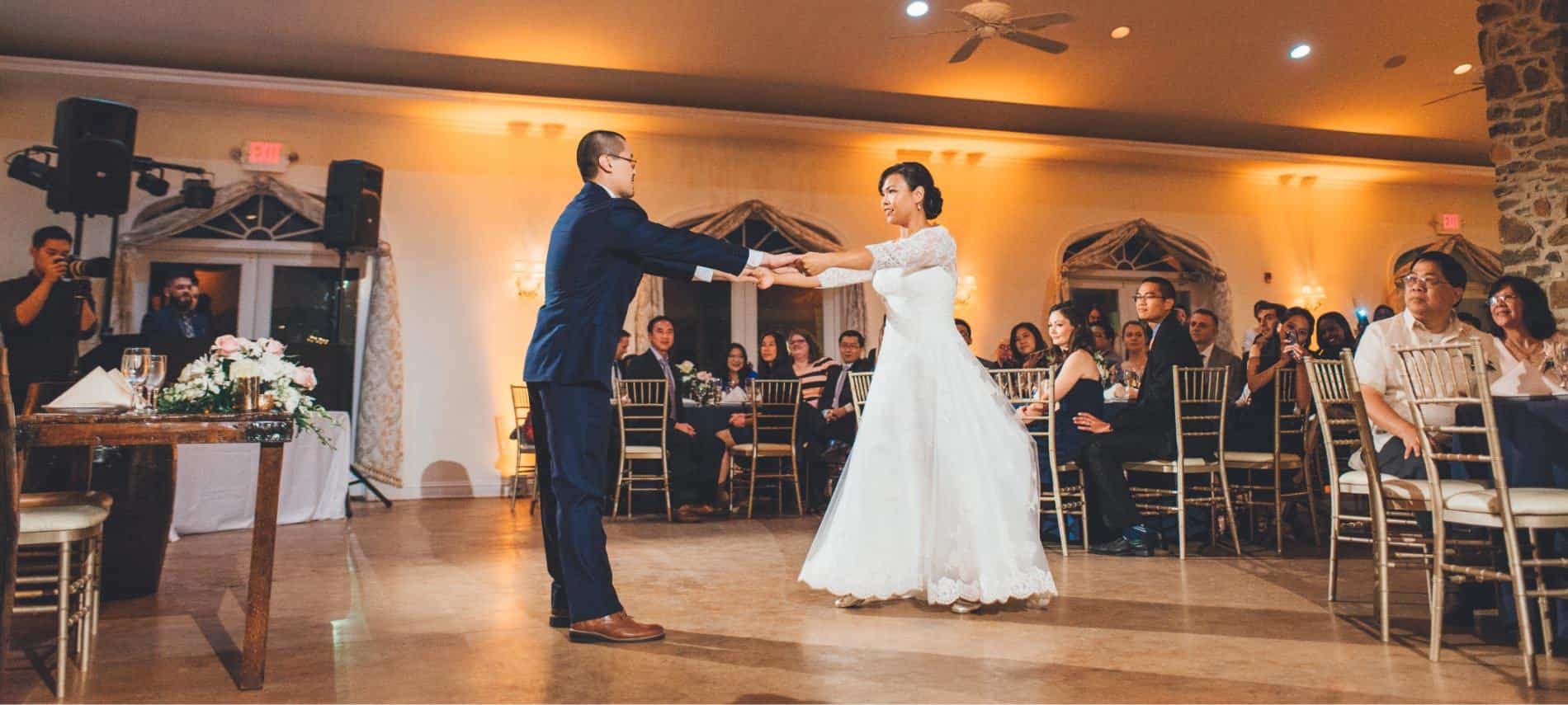 Bride in white dress and groom in dark blue suit dancing with people in the background watching the couple