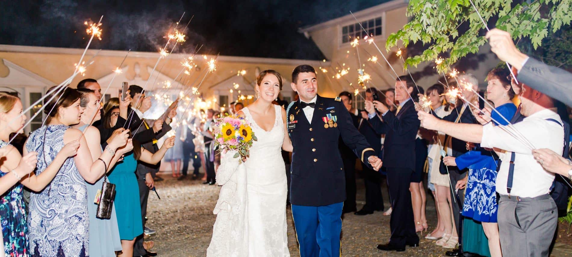 Bride with white wedding dress and groom in uniform walking through people holding sparklers