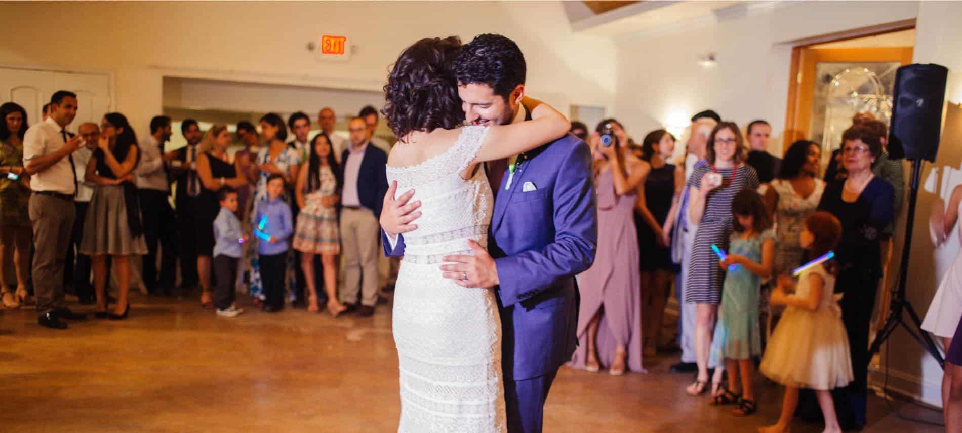 Bride in white dress and groom in dark purple suit dancing with people in the background watching the couple