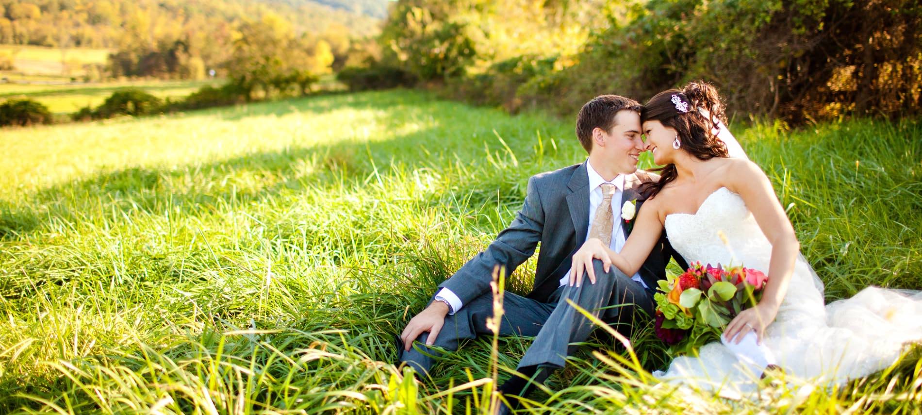 Bride in white wedding dress and groom in dark gray suit sitting together surrounded by green grass