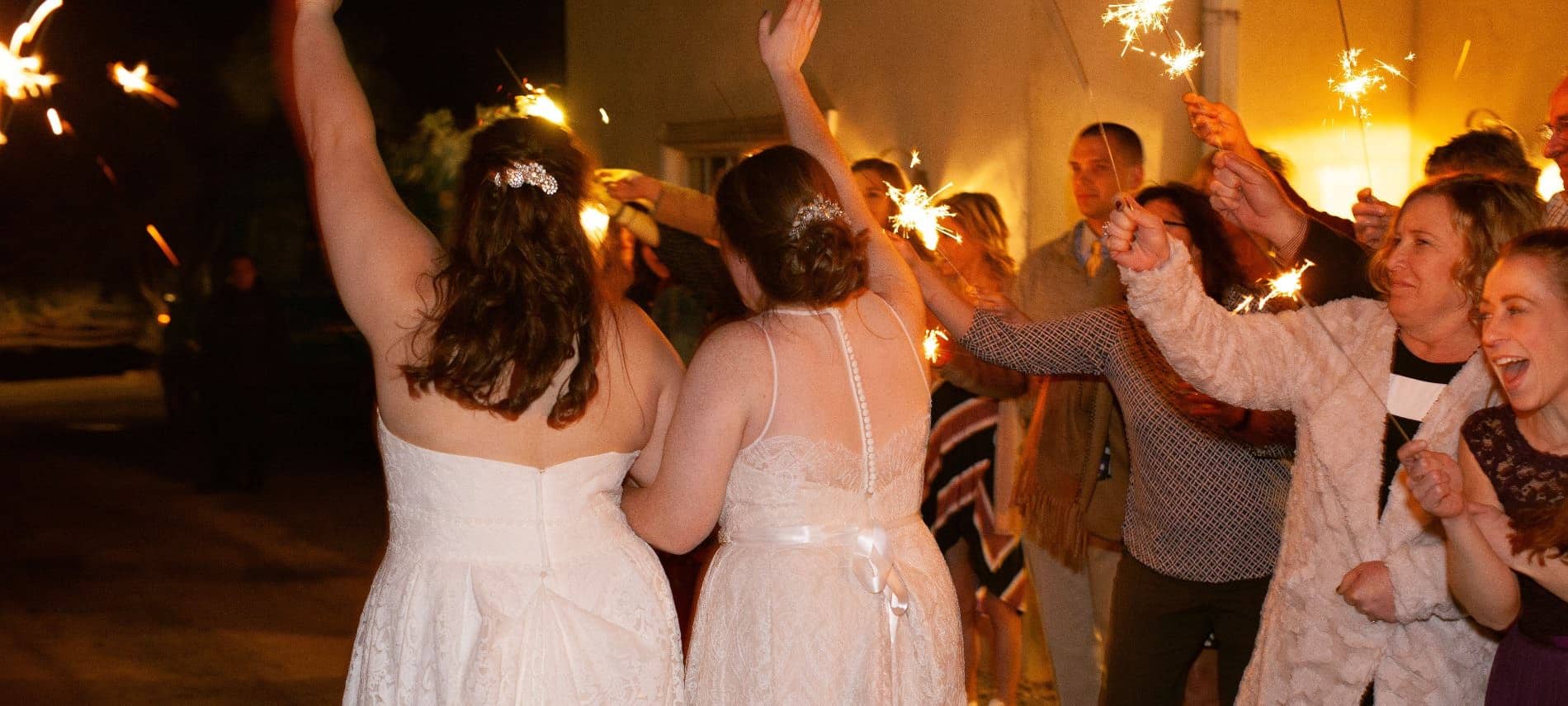 Two brides with white wedding dresses walking through people holding sparklers