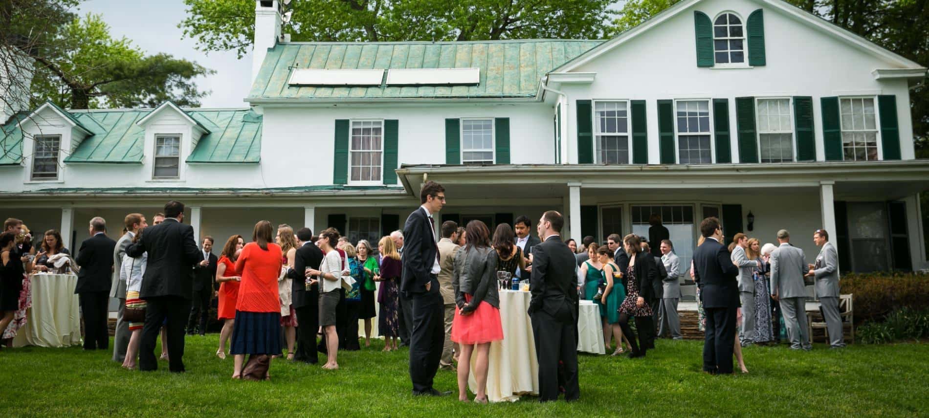Exterior view of a white house with dark green shutters and roof and wedding guests standing on the green lawn for a cocktail reception
