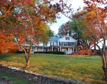 Exterior view of property painted white with dark shutters surrounded by a large green lawn and trees with fall color