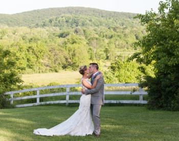 Bride in white dress and groom in gray suit kissing with white fence, green trees, and rolling hills in the background