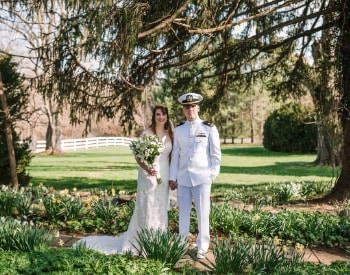 Bride with white dress and groom with white naval uniform standing on path between flowers