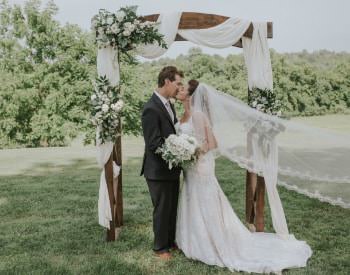 Bride in white dress with long train and groom with dark gray suit kissing under an alter draped in white fabric and white flowers